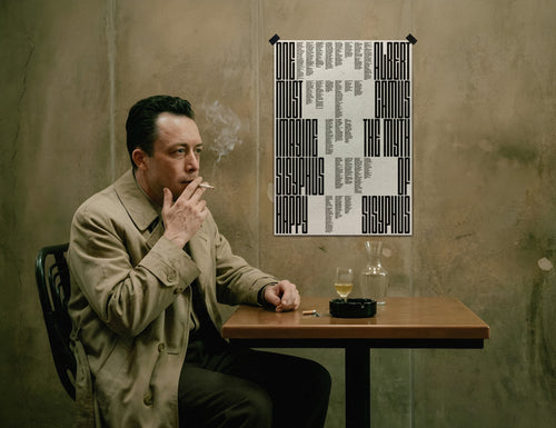 Albert Camus in a beige trench coat sits at a wooden table smoking, with a framed black-and-white typographic the Myth of sisyphus poster print on the wall behind him and a glass and ashtray on the table.
